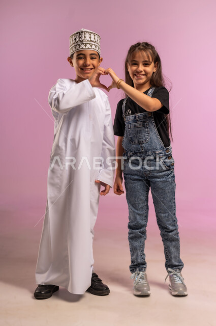 العلاقة الاخوية الايجابية، مفهوم الحب والمودة بين الاصدقاء، بورتريه لولد عربي خليجي عماني يرتدي الدشداشة والكمة يقوم بتشكيل قلب بيده مع اخته ينظر إلى الكاميرا بتعابير الحب والألفة والسعادة, صورة الجسم بالطول الكامل، خلفية وردية