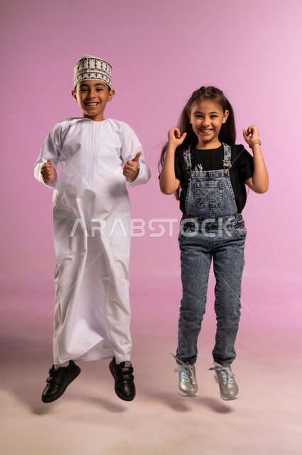 القفز والنظر الى الكاميرا بتعابير السعادة والحماس والسرور، مفهوم الانجاز وتحقيق الهدف، بورتريه لولد عربي خليجي عماني مبتسم يرتدي الدشداشة والكمة يقف بجانب اخته يرفعان قبضة يديهما للاعلى بإيماءات الفوز والانتصار والنجاح, الجسم بالطول الكامل، خلفية وردية