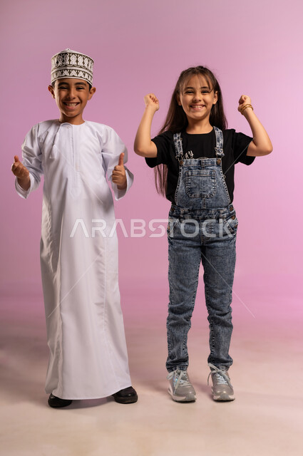 مفهوم الانجاز وتحقيق الهدف، بورتريه لولد عربي خليجي عماني مبتسم يرتدي الدشداشة والكمة يقف بجانب اخته يرفعان قبضة يديهما للاعلى بإيماءات الفوز والانتصار والنجاح, النظر الى الكاميرا بتعابير السعادة والقوة والسرور، الجسم بالطول الكامل، خلفية وردية