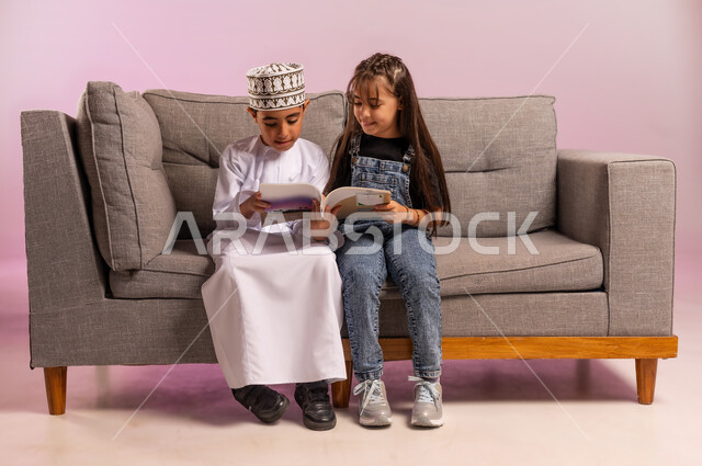 الاهتمام بتصفح الكتب المفيدة, بورتريه لولد عربي خليجي عماني يرتدي الدشداشة والكمة يجلس على كنبه مريحة يمسك بيده كتاب برفقة أخته, قضاء وقت الفراغ بالتثقيف والمطالعة مع الأصدقاء, تنمية مهارة القراءة, مفهوم الثقافة والتعلم الذاتي، خلفية وردية
