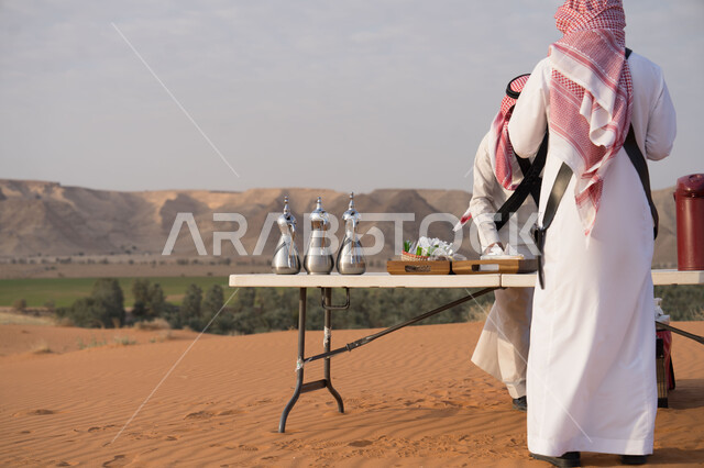The concept of hospitality and good reception, a back view of a Saudi Arabian Gulf man wearing a traditional shemagh and thobe standing in the desert in front of a table with metal coffee pots on it, authentic customs and traditions in the Kingdom of Saudi Arabia