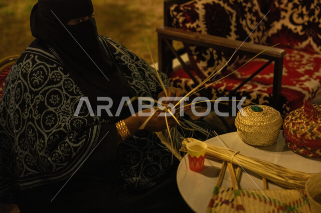 الأصالة والتراث في السعودية, حرف يدوية قديمة تاريخية, مهن نسائية شعبية تقليدية, استخدام أوراق النخيل المجففة في صناعة السلل, منتجات محلية وطنية, صورة مقربة من الجانب لامرأة عربية خليجية سعودية محجبة ترتدي الخمار تقوم بجدل سعائف الخوص