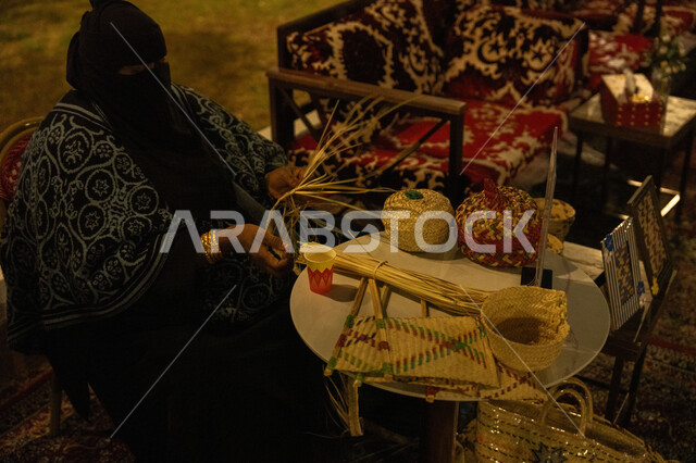 استخدام أوراق النخيل المجففة في صناعة السلل, الأصالة والتراث في السعودية, حرف يدوية قديمة تاريخية, مهن نسائية شعبية تقليدية, منتجات محلية وطنية, صورة من الجانب لامرأة عربية خليجية سعودية محجبة ترتدي الخمار تقوم بجدل سعائف الخوص