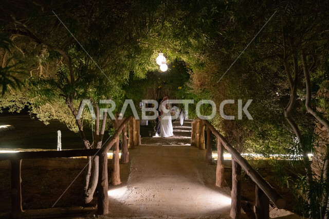 Enjoying the scenery of green trees and plants, walking in a park in Saudi Arabia, a Saudi Arabian Gulf man wearing a traditional shemagh and thobe holding a metal coffee pot walking in a public park, serving hot drinks