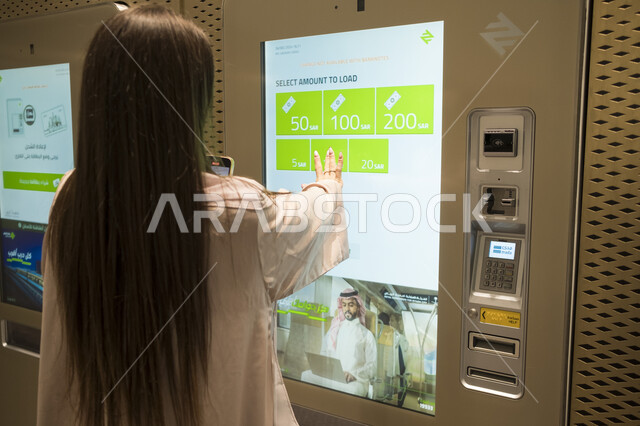 تسهيل الحركة المرورية للشعب السعودي, تطور وتقدم وسائل النقل والمواصلات في المملكة العربية السعودية, مفهوم التنقل السريع والآمن, صورة مقربة من الخلف لامرأة عربية خليجية سعودية ترتدي الزي الكاجوال تقوم بالدفع في ماكينة التذاكر في محطة مترو الرياض