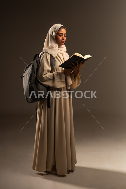 تعابير النشاط والاجتهاد، الاستعداد للعام الدراسي الجديد, التعليم العالي في الجامعات الأكاديمية في السعودية, بورتريه لطالبة جامعية عربية خليجية سعودية محجبة ترتدي العباءة وتضع حقيبة الظهر تحمل بيدها كتاب مفتوح, صورة الجسم بالطول الكامل، خلفية رمادية