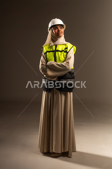 مفهوم الهندسة والإنشاء المعماري، مهنة ووظيفة نسائية، بورتريه لمهندسة عربية خليجية سعودية محجبة ترتدي العباءة والسترة و خوذة الحماية تقف بأيدي متقاطعة تنظر الى شيء ما في الاعلى بإيماءات الثقة بالنفس، صورة الجسم بالطول الكامل، خلفية رمادية