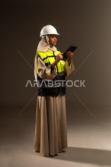 مراجعة المخططات عبر الجهاز اللوحي، مفهوم الهندسة والإنشاء المعماري, تصفح مواقع التواصل الاجتماعي، بورتريه لمهندسة عربية خليجية سعودية محجبة ترتدي العباءة والسترة و خوذة الحماية تمسك بيدها التابليت، صورة الجسم بالكامل، خلفية رمادية