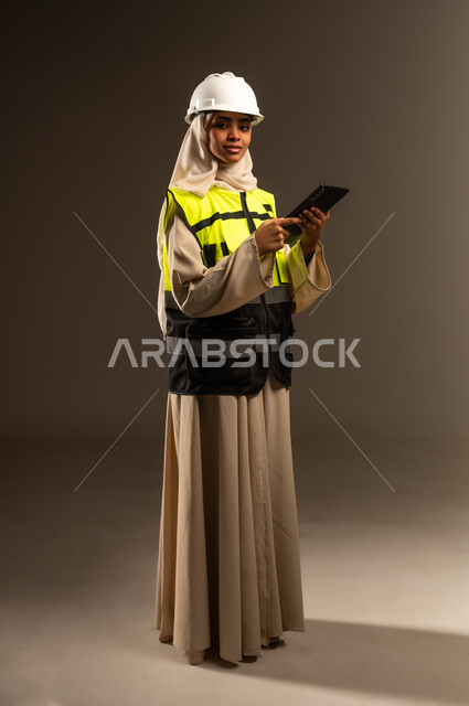 النظر الى الكاميرا بإيماءات السرور، مراجعة المخططات عبر الجهاز اللوحي، مفهوم الهندسة والإنشاء المعماري, بورتريه لمهندسة عربية خليجية سعودية محجبة ترتدي العباءة والسترة و خوذة الحماية تمسك بيدها التابلت، صورة الجسم بالطول الكامل، خلفية رمادية