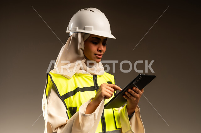 مراجعة المخططات عبر الجهاز اللوحي، مفهوم الهندسة والإنشاء المعماري, تصفح مواقع التواصل الاجتماعي، استخدام جهاز تقني حديث متطور، بورتريه مقرب لمهندسة عربية خليجية سعودية محجبة ترتدي العباءة والسترة و خوذة الحماية تمسك بيدها جهاز التابليت المحمول، خلفية رمادية