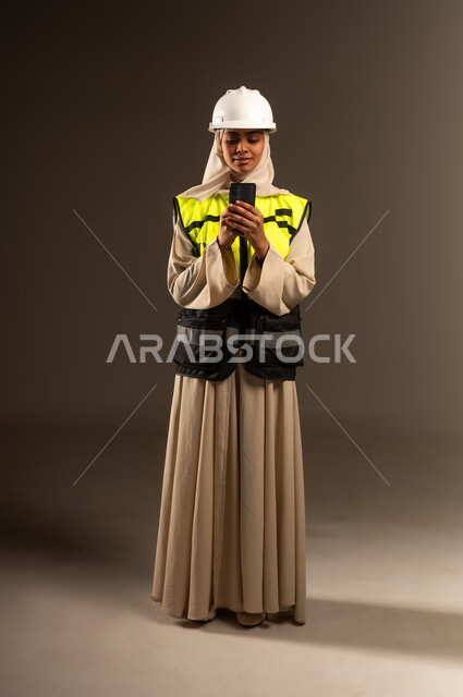 مفهوم الهندسة والإنشاء المعماري, الدردشة مع العائلة والأصدقاء، تصفح مواقع التواصل الاجتماعي، استخدام جهاز تقني حديث متطور، بورتريه لمهندسة عربية خليجية سعودية محجبة ترتدي العباءة والسترة و خوذة الحماية تمسك بيدها الهاتف المحمول، صورة الجسم بالطول الكامل، خلفية رمادية