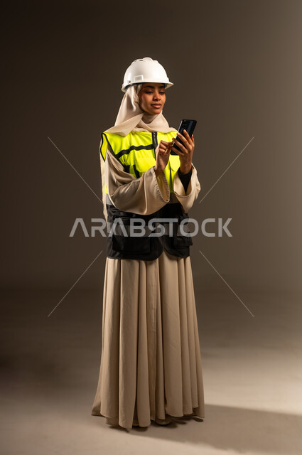 Chatting with family and friends, browsing social media, using a modern advanced technical device, concept of engineering and architectural construction, portrait of a veiled Saudi Arabian Gulf female engineer wearing an abaya, jacket and protective helmet holding a mobile phone in her hand, full body image, gray background