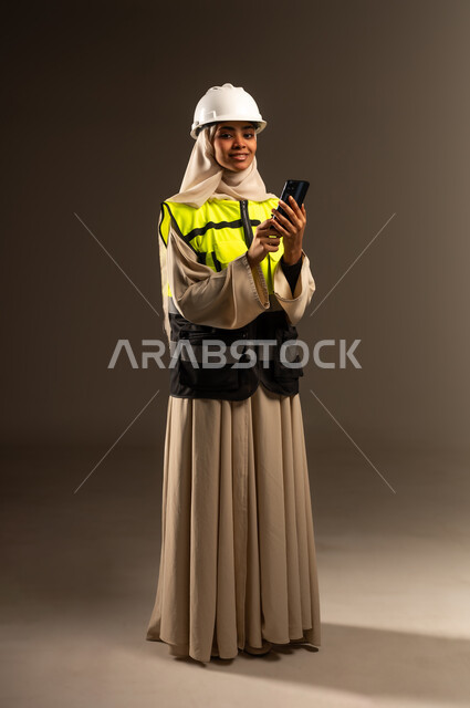 تصفح مواقع التواصل الاجتماعي، استخدام جهاز تقني حديث متطور، مفهوم الهندسة والإنشاء المعماري, بورتريه لمهندسة عربية خليجية سعودية محجبة ترتدي العباءة والسترة و خوذة الحماية تنظر الى الكاميرا تمسك بيدها الهاتف المحمول، صورة الجسم بالطول الكامل، خلفية رمادية