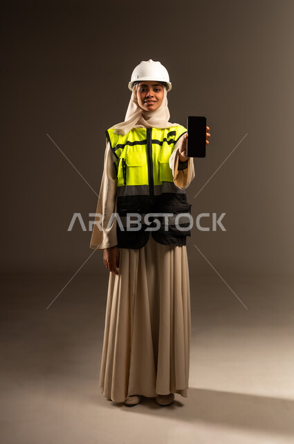 عرض الهاتف المحمول بشاشة سوداء فارغة، مفهوم الهندسة والإنشاء المعماري, بورتريه لمهندسة عربية خليجية سعودية محجبة ترتدي العباءة والسترة و خوذة الحماية تنظر الى الكاميرا تمسك بيدها الجوال، استخدام جهاز تقني حديث متطور، صورة الجسم بالطول الكامل، خلفية رمادية
