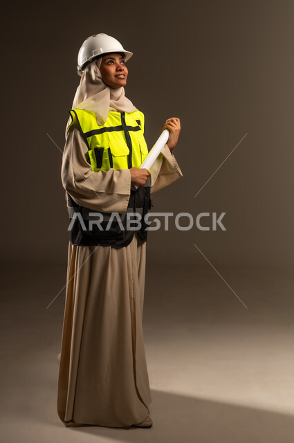 مفهوم الهندسة والإنشاء المعماري، التحقق من تنفيذ المشروع الإنشائي, مهنة ووظيفة نسائية، بورتريه من الجانب لمهندسة عربية خليجية سعودية محجبة ترتدي العباءة والسترة و خوذة الحماية تنظر الى شيء ما في الاعلى تمسك بيدها المخطط الهندسي، صورة الجسم بالكامل، خلفية رمادية