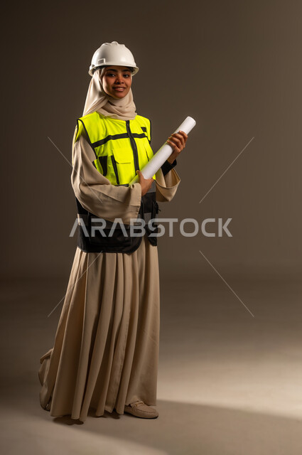 Looking at the camera with gestures of self-confidence, concept of engineering and architectural construction, checking the implementation of the construction project, portrait of a veiled Saudi Arabian Gulf female engineer wearing an abaya, jacket and protective helmet holding an engineering drawing in her hand, full-length body image, gray background