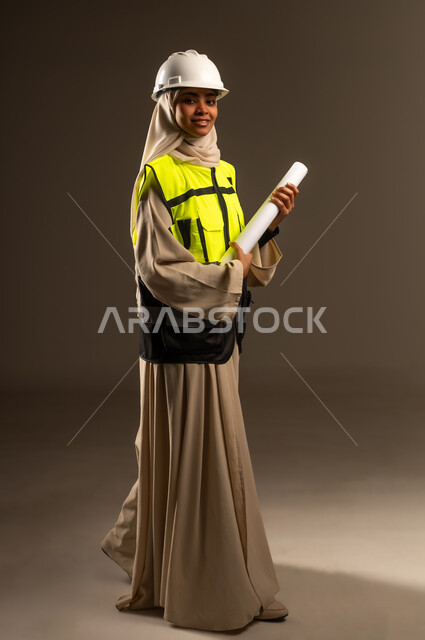 مهنة ووظيفة نسائية، مفهوم الهندسة والإنشاء المعماري، التحقق من تنفيذ المشروع الإنشائي, بورتريه لمهندسة عربية خليجية سعودية محجبة ترتدي العباءة والسترة و خوذة الحماية تنظر الى الكاميرا تمسك بيدها المخطط الهندسي، صورة الجسم بالكامل، خلفية رمادية
