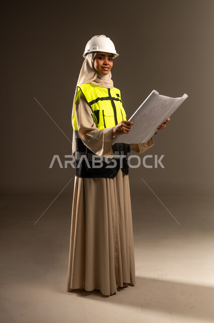 Looking at the camera with gestures of self-confidence, concept of engineering and architectural construction, checking the implementation of the construction project, portrait of a veiled Saudi Arabian Gulf female engineer wearing an abaya, jacket and protective helmet holding an engineering drawing in her hand, full-length body image, gray background