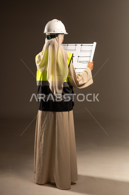 Concept of engineering and architectural construction, verification of construction project implementation, Saudi female profession and job, portrait from the back of a veiled Saudi Arabian Gulf female engineer wearing an abaya, jacket and protective helmet holding an engineering plan in her hand, full-length body image, gray background
