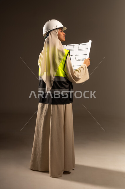 Verifying the implementation of the construction project, Saudi female profession and job, concept of engineering and architectural construction, portrait from the back of a veiled Saudi Arabian Gulf female engineer wearing an abaya, jacket and protective helmet holding an engineering plan in her hand, full-length body image, gray background