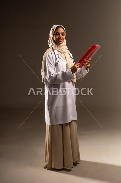 النظر الى الكاميرا بإيماءات السرور، مهنة ووظيفة نسائية، مراجعة ملفات المرضى، العمل في القطاع الصحي في المملكة، بورتريه لطبيبة عربية خليجية سعودية محجبة ترتدي العباءة و المعطف الابيض تمسك بيدها ملف المريض، صورة الجسم بالطول الكامل، خلفية رمادية