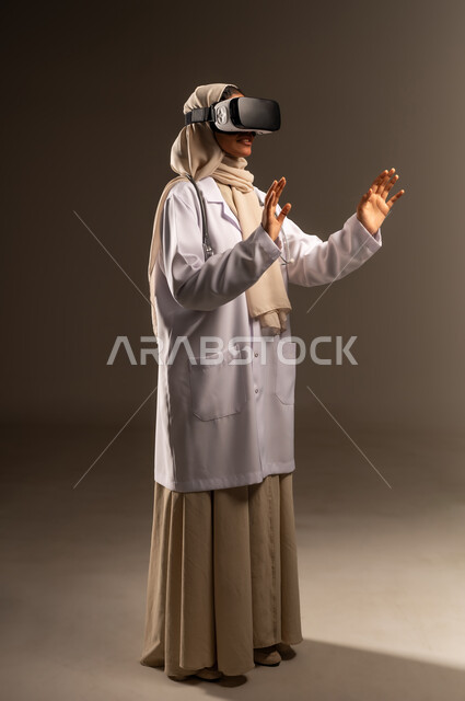 مشاهدة افلام الفي آر ثلاثي الابعاد، التفاعل مع العناصر الهولوجرامية، بورتريه لطبيبة عربية خليجية سعودية محجبة ترتدي العباءة والمعطف الأبيض ونظارات الواقع الإفتراضي في آر ترفع يديها للاعلى تحاول لمس عنصر ما، صورة الجسم بالكامل، خلفية رمادية