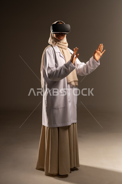 التفاعل مع العناصر الهولوجرامية، بورتريه لطبيبة عربية خليجية سعودية محجبة ترتدي العباءة والمعطف الأبيض ونظارات الواقع الإفتراضي في آر ترفع يديها للاعلى تحاول لمس عنصر ما، مشاهدة افلام الفي آر ثلاثي الابعاد، صورة الجسم بالكامل، خلفية رمادية