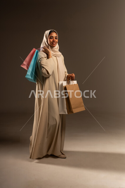 الاثر الايجابي للتسوق على نفسية النساء، الاستفادة من العروض والخصومات, شراء المستلزمات والهدايا, بورتريه لإمرأة عربية خليجية سعودية محجبة ترتدي العباءة تحمل بيدها أكياس كرتون ملونه تنظر الى الكاميرا بإيماءات الفرح والسرور، صورة الجسم بالطول الكامل, خلفية رمادية