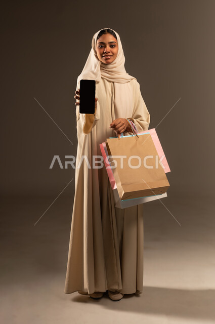 عرض شاشة سوداء فارغة عبر الهاتف المحمول, الاثر الايجابي للتسوق على نفسية النساء، الاستفادة من العروض والخصومات, بورتريه لإمرأة عربية خليجية سعودية مبتسمة محجبة ترتدي العباءة تنظر الى الكاميرا تحمل بيدها الجوال و أكياس كرتون ملونه، صورة الجسم بالكامل, خلفية رمادية