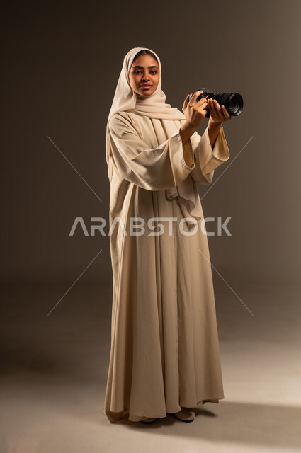 احتراف فن التصوير الفوتوغرافي باستخدام كاميرات رقمية عالية الدقة, وظيفة ومهنة نسائية سعودية, بورتريه لإمراة عربية خليجية سعودية محجبة ترتدي العباءة تنظر الى الكاميرا بإيماءات السرور، التقاط الصور التذكارية عبر كاميرا فوتوغرافية, صورة الجسم بالطول الكامل، خلفية رمادية