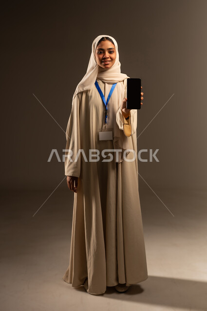 موك اب ابيض، النظر الى الكاميرا بتعابير السرور, عرض شاشه سوداء فارغه عبر الهاتف المحمول, استخدام جهاز تقني حديث متطور, بورتريه لامرأة عربية خليجية سعودية محجبة ترتدي العباءة تضع بطاقة بيضاء فارغه بشريط أزرق حول رقبتها، الجسم بالطول الكامل, خلفيه رمادية