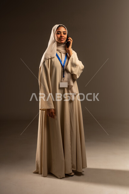 التواصل مع الاهل والاصدقاء، اجراء مكالمه هاتفية عبر الهاتف المحمول, استخدام جهاز تقني حديث متطور, بورتريه لامرأة عربية خليجية سعودية محجبة ترتدي العباءة تضع بطاقة بيضاء فارغه بشريط أزرق حول رقبتها، موك اب ابيض، صورة الجسم بالطول الكامل, خلفيه رمادية