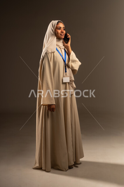 اجراء مكالمه هاتفية عبر الهاتف المحمول, استخدام جهاز تقني حديث متطور, بورتريه لامرأة عربية خليجية سعودية محجبة ترتدي العباءة تضع بطاقة بيضاء فارغه بشريط أزرق حول رقبتها، موك اب ابيض، التواصل مع الاهل والاصدقاء، صورة الجسم بالطول الكامل, خلفيه رمادية