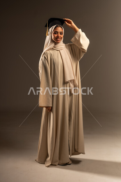 النظر الى الكاميرا بإيماءات الفخر والسرور, الاحتفالات و المناسبات  الجامعية, الفرح بتحقيق حلم النجاح ,بورتريه لطالبة عربية سعودية خليجية محجبة مبتسمة ترتدي العباءة تمسك بيدها قبعه التخرج, صورة الجسم بالطول الكامل, خلفية رمادية
