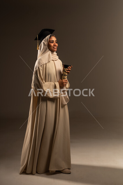 المشي و الذهاب للتكريم بتعابير الحماس, الفرح بتحقيق حلم النجاح, المناسبات والاحتفالات الجامعية, النظر الى شيء ما بإيماءات الفخر والسرور ,بورتريه لطالبة عربية سعودية خليجية ترتدي العباءة والقبعه تحمل بيدها وثيقة التخرج, صورة الجسم بالطول الكامل, خلفية رمادية