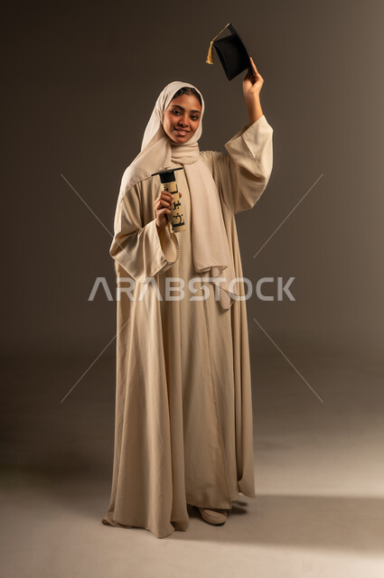 المناسبات والاحتفالات الجامعية, النظر الى الكاميرا بإيماءات الفخر والسرور ,الفرح بتحقيق حلم النجاح, بورتريه لطالبة عربية سعودية خليجية ترتدي العباءة تمسك بيدها القبعه ووثيقة التخرج بتعابير الحماس, صورة الجسم بالطول الكامل, خلفية رمادية