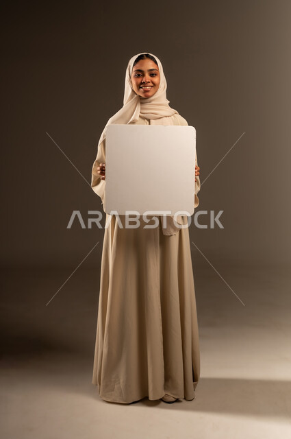 موك اب مربع أبيض, النظر الى الكاميرا بتعابير السرور, العروض والإعلانات التسويقية, بورتريه لإمرأة عربية خليجية سعودية محجبة ترتدي العباءة تحمل بيديها لوحة بيضاء مربعة فارغة, لوحات توضيحية ووسائل تعليمية, الطول الكامل للجسم, خلفية رمادية