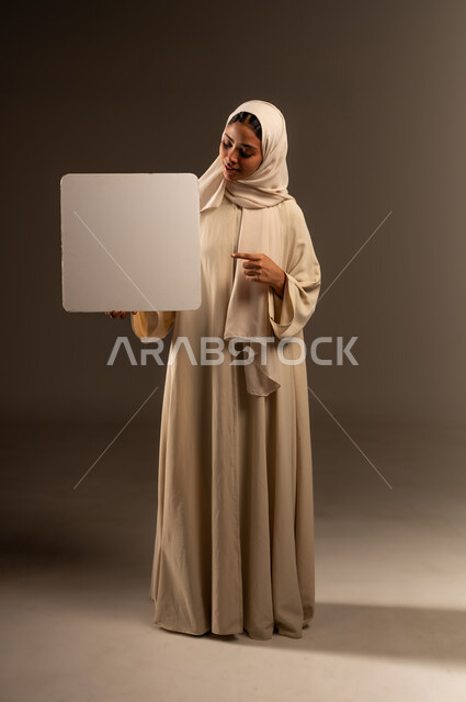 Pointing index finger at white square mockup, marketing offers and advertisements, portrait of a veiled Saudi Arabian Gulf woman wearing an abaya holding a blank white square board in her hand, illustrations and educational tools, full body length, gray background