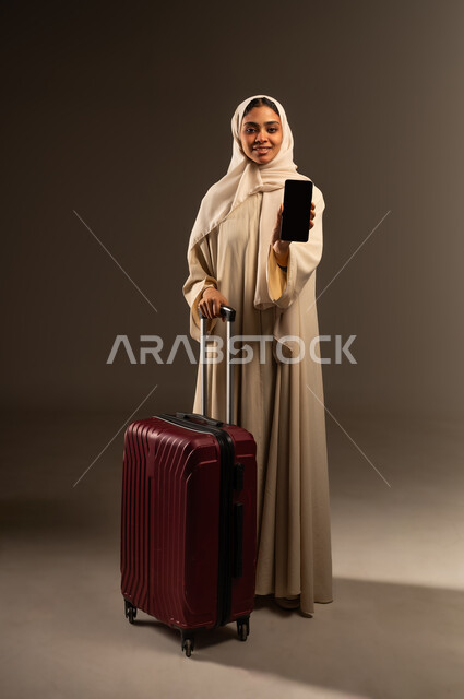 Having a pleasant trip abroad, using a modern advanced technical device, portrait of a veiled Saudi Arabian Gulf woman wearing an abaya holding a travel bag and looking at the camera, displaying a blank black screen via mobile phone, full-length body image, gray background