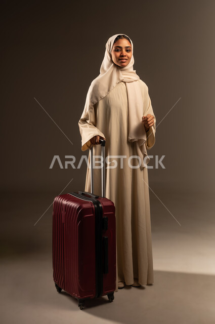 قضاء رحلة صيفية ممتعة, الاستعداد لبدء العطلة خارج البلاد ,السياحة في إجازة الصيف, بورتريه لشابة عربية خليجية سعودية محجبة ترتدي العباءة تمسك بيدها حقيبة السفر تنظر إلى الكاميرا بإيماءات السرور، صورة الجسم بالطول الكامل, خلفية رمادية
