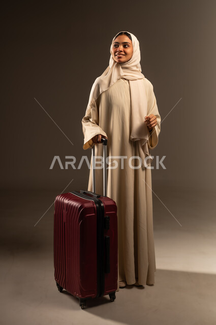 الاستعداد لبدء العطلة خارج البلاد ,السياحة في إجازة الصيف, قضاء رحلة صيفية ممتعة, بورتريه لشابة عربية خليجية سعودية محجبة ترتدي العباءة تمسك بيدها حقيبة السفر وتنظر إلى شيء ما بإيماءات السرور، صورة الجسم بالطول الكامل, خلفية رمادية