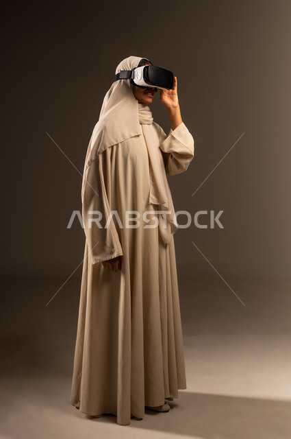 استخدام تقنية الكترونية حديثة، الاندماج ومحاكاة العالم الخيالي، بورتريه لإمراة عربية خليجية سعودية محجبة ترتدي العباءة تمسك بيدها نظارات ثلاثية الابعاد (الفي ار)، المتعة والتسلية بالمشاهدة عبر نظارة الواقع الافتراضي، صورة الجسم بالطول الكامل، خلفية رمادية