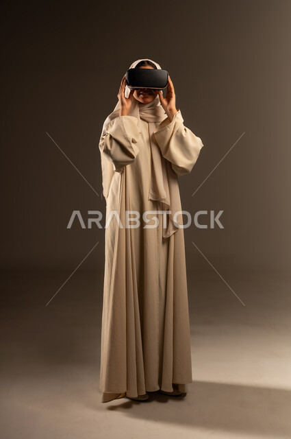 الاندماج ومحاكاة العالم الخيالي، استخدام تقنية الكترونية حديثة، بورتريه لإمراة عربية خليجية سعودية محجبة ترتدي العباءة تمسك بيديها نظارات ثلاثية الابعاد (الفي ار)، المتعة والتسلية بالمشاهدة عبر نظارة الواقع الافتراضي، صورة الجسم بالطول الكامل، خلفية رمادية