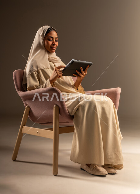 إدارة وتنظيم العمل عن بعد، مفهوم انجاز المهام اليومية، استخدام جهاز تقني حديث تكنولوجي متطور، بورتريه لامرأة عربيه خليجيه سعوديه محجبه مبتسمة ترتدي العباءة تحمل بيدها التابلت بتعابير السرور، خلفية رمادية