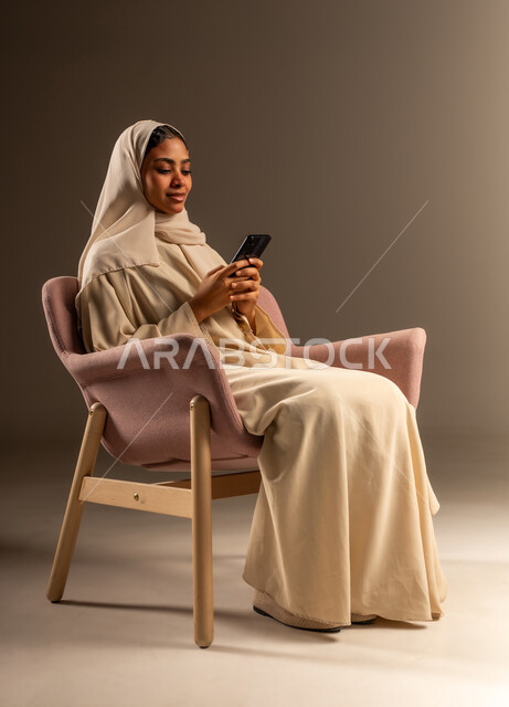 قضاء وقت ممتع عبر الجوال مع الأصحاب، تصفح مواقع التواصل الاجتماعي، بورتريه لامرأة عربية خليجية سعودية محجبة ترتدي العباءة تجلس على كرسي مريح تقوم بإرسال الرسائل النصية عبر الهاتف المحمول، خلفية رمادية