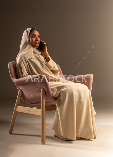 استخدام جهاز تقني حديث و متطور, بورتريه لامرأة عربية خليجية سعودية محجبة ترتدي العباءة تجلس على كرسي مريح تقوم بإجراء مكالمة صوتية عبر الهاتف المحمول، الدردشة والتواصل مع العائلة والأصدقاء، خلفية رمادية