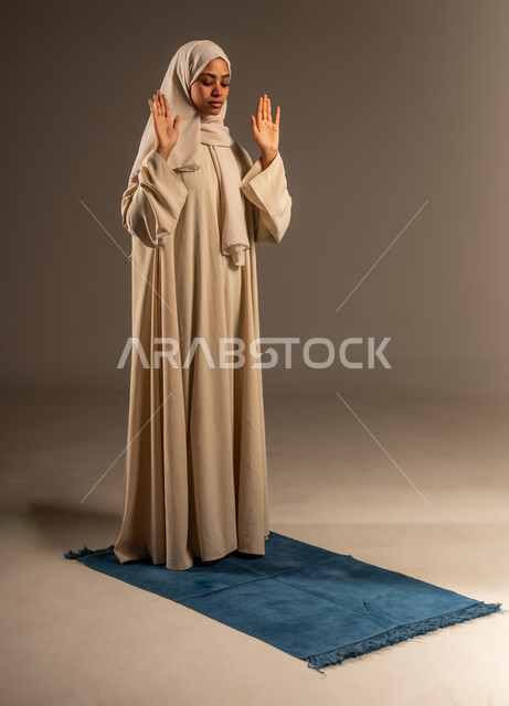 الحفاظ على إقامة الصلوات الخمس بوقتها، العبادة و التقرب الى الله تعالى في شهر رمضان، بورتريه لإمرأة عربية سعودية خليجية محجبة ترتدي العباءة تقف على السجادة ترفع يديها للاعلى تقوم بآداء فريضة الصلاة، صورة الجسم بالطول الكامل، خلفية رمادية
