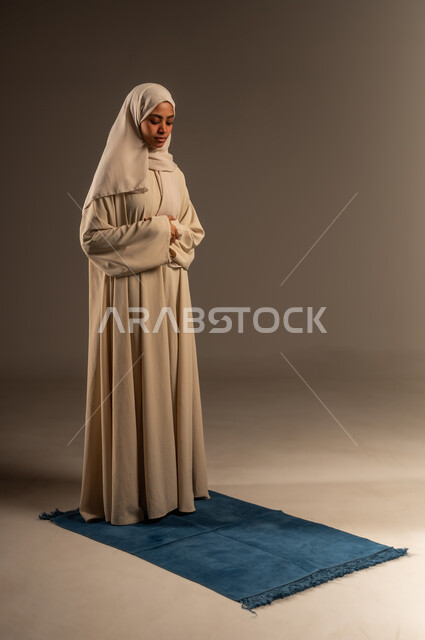 العبادة و التقرب الى الله تعالى في شهر رمضان، الحفاظ على إقامة الصلوات الخمس بوقتها، بورتريه لإمرأة عربية سعودية خليجية محجبة ترتدي العباءة تقف على السجادة تقوم بآداء فريضة الصلاة، صورة الجسم بالطول الكامل، خلفية رمادية