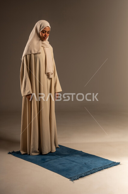 الحفاظ على إقامة الصلوات الخمس بوقتها، مفهوم العبادة و التقرب الى الله تعالى في شهر رمضان، بورتريه لإمرأة عربية سعودية خليجية محجبة ترتدي العباءة تقف على السجادة تقوم بآداء فريضة الصلاة، صورة الجسم بالطول الكامل، خلفية رمادية
