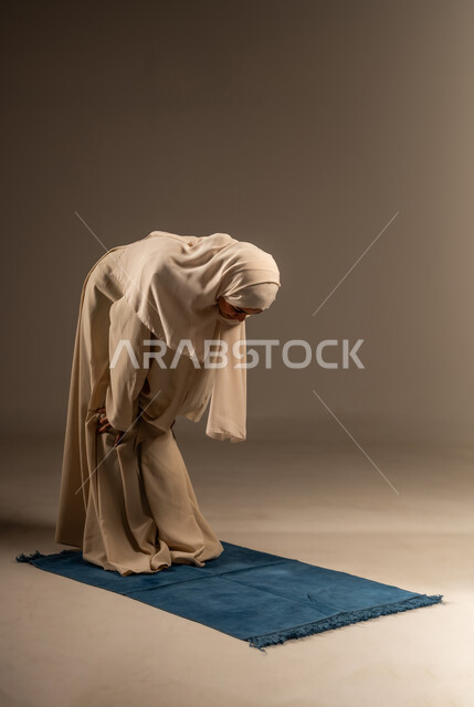 The concept of worship and drawing closer to God Almighty, striving in acts of obedience, supplications and remembrances, maintaining the five daily prayers on time, a portrait of a veiled Saudi Arabian Gulf woman wearing an abaya standing on a prayer rug, kneeling while performing the obligatory prayer, gray background